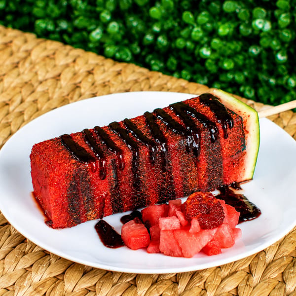 Watermelon on a stick, drizzled with dark sauce and chili powder, served with diced watermelon.
