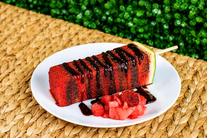 Watermelon on a stick, drizzled with dark sauce and chili powder, served with diced watermelon.