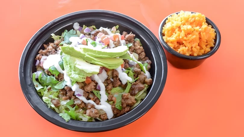 Taco salad bowl with ground beef, lettuce, avocado, and sour cream, with a side of rice.