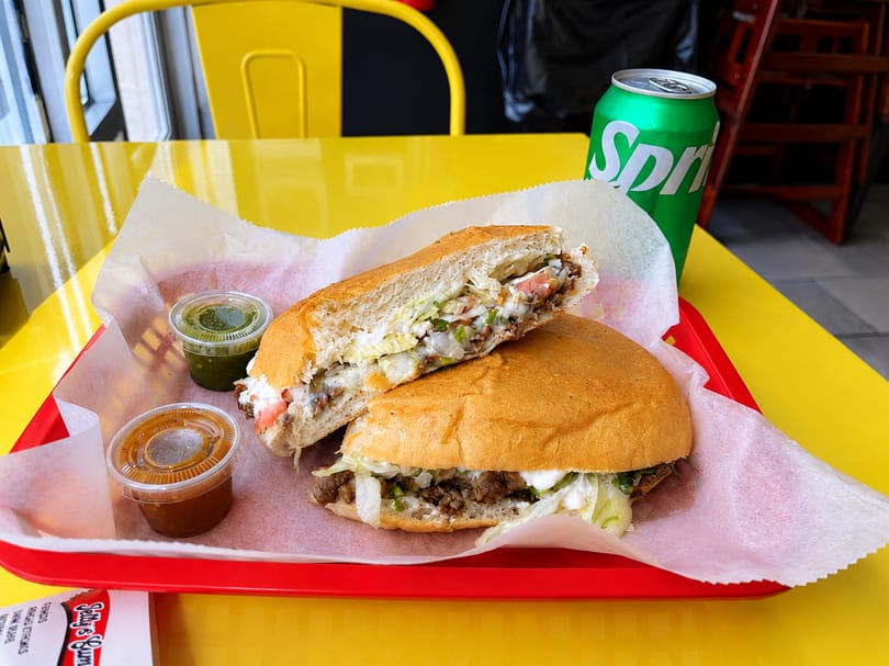 A torta sandwich, cut in half, with two sauces and a Sprite on a yellow table.
