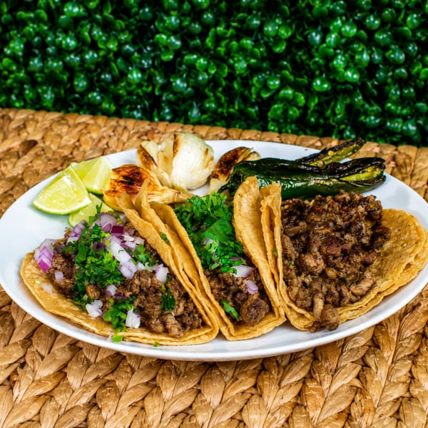 Three street tacos with meat, cilantro, and onions, served with grilled peppers, garlic, and lime.