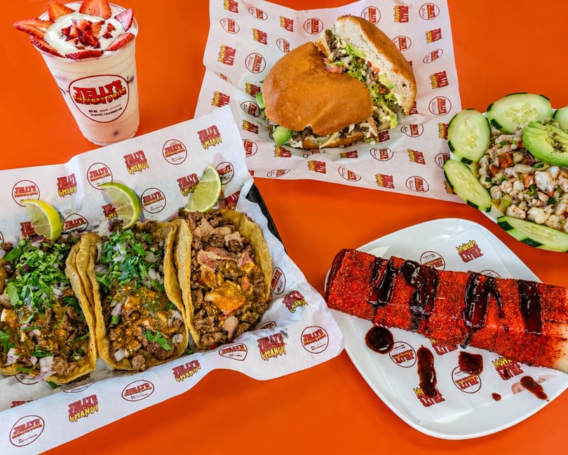 A vibrant orange table laden with Mexican dishes: tacos, a torta, ceviche, a strawberry drink, and a red dessert.