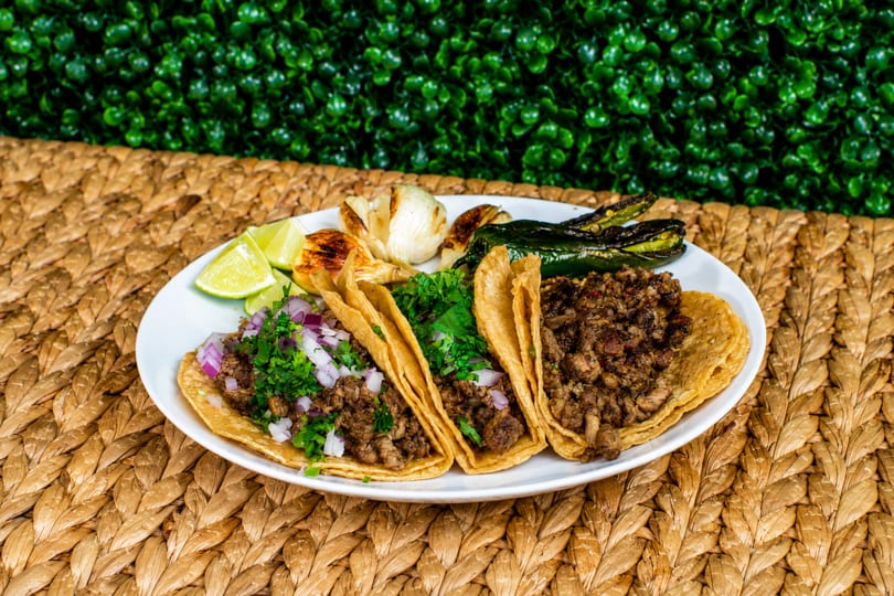 Three street tacos with meat, cilantro, and onions, served with grilled peppers, garlic, and lime.
